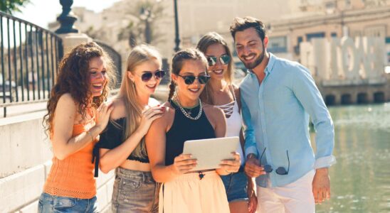 Five friends laughing together by the waterfront in Malta, enjoying a sunny day and sharing a tablet.