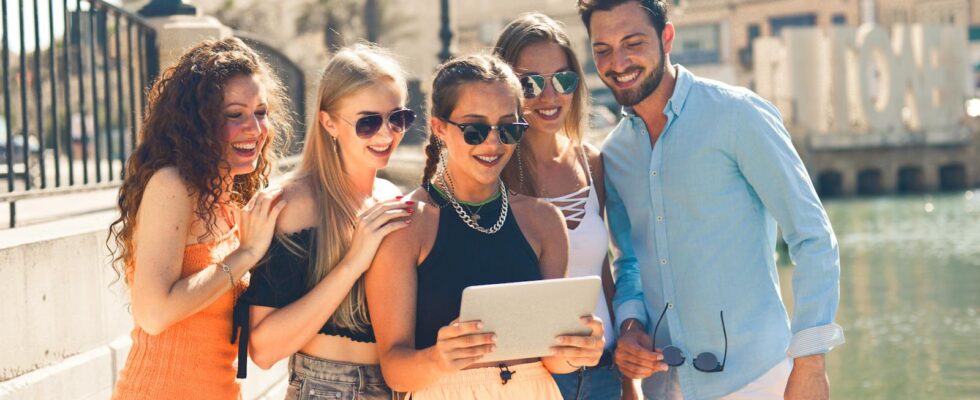 Five friends laughing together by the waterfront in Malta, enjoying a sunny day and sharing a tablet.